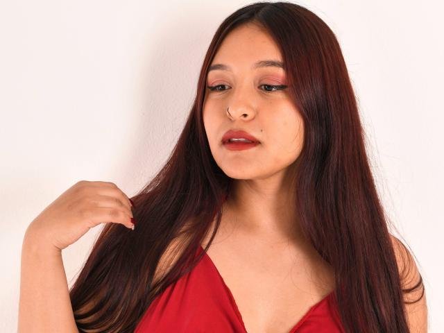 MarianaVargasX with long red hair, red lipstick, and a nose piercing, wearing a red dress, looking down, touching her hair with her right hand.