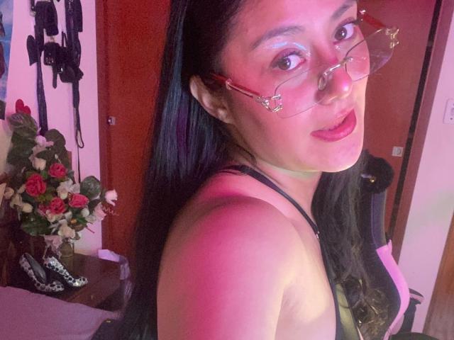 CristalZafiro, con gafas, lápiz labial rosado-rojizo y cabello largo y negro, mostrando su hombro descubierto y una camiseta negra, sonriendo a la cámara. Sus senos son parcialmente visibles..