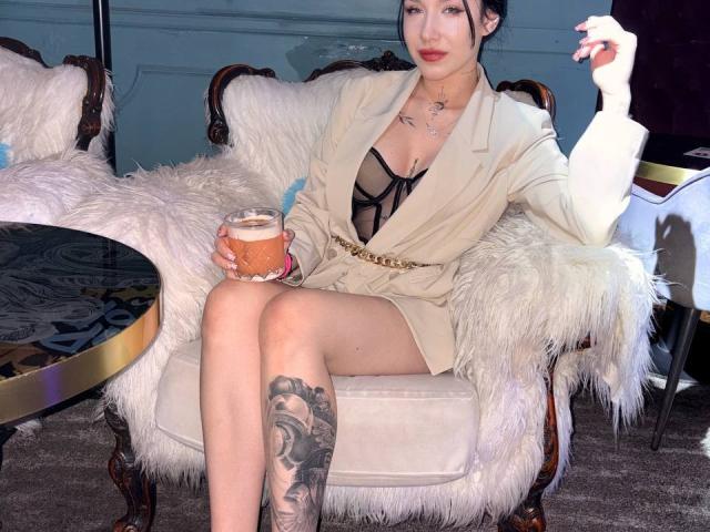 NikkiGoldX, in einem beigen Kleid, sitzt auf einem flauschigen Stuhl, hält eine Tasse Kaffee in der Hand, hat ein Tattoo an ihrem rechten Oberschenkel und trägt einen selbstbewussten Gesichtsausdruck, wobei sie ihre linke Hand hebt..