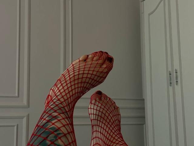NikkiGoldX's feet in red fishnet socks, toes pointing upward, with visible toenails, legs crossed at the ankles, in a room with white cabinets.