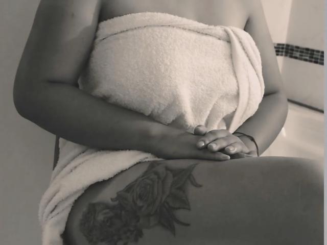 CristalWil with a towel covering her tits, hands clasped, showing a detailed rose tattoo on her right thigh, in black-and-white.