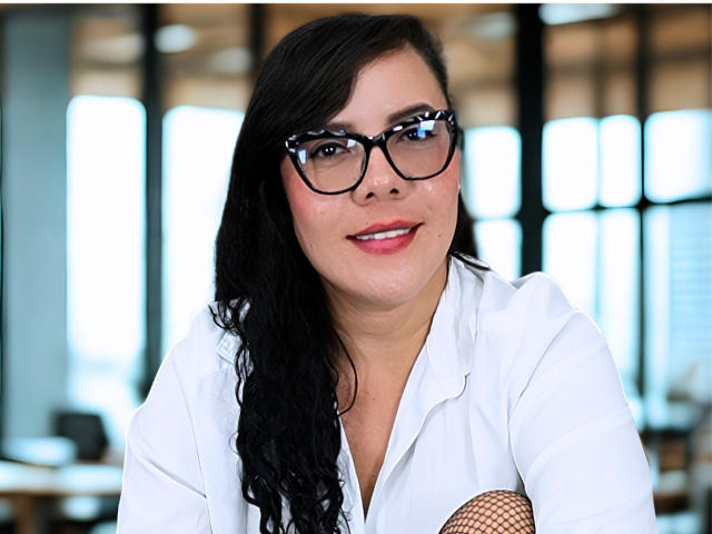 Une femme latina attrayante, avec des cheveux noirs ondulés, des lunettes, une chemise blanche et une manche en résille, souriante, montrant ses dents, avec du rouge à lèvres rose, dans une pièce bien éclairée..