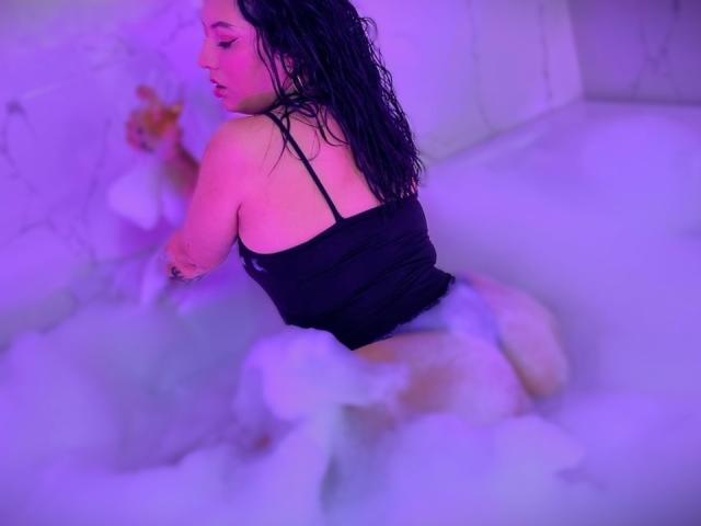 AmyRoss with wet black hair, wearing a black tank top, kneels in a bubbly white bath, her butt raised, surrounded by purple-lit foam, looking over her shoulder.