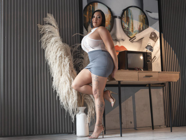 MellissaCortez with large tits, curvy ass, and tattoos, wearing a sheer white top and grey skirt, leaning on a table with puffy grass in front of a retro TV.