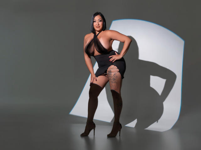 MellissaCortez with big boobs in a sheer black top, tight black skirt, thigh-high brown boots, and a tattoo on her right thigh, standing confidently, shadow on the wall.