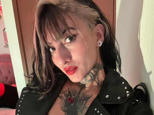 LiaMiss with black and blonde hair, red lipstick, nose ring, tattoos, white ear gauges, black spiked leather jacket, and chest tattoo, looking at camera, in bedroom.