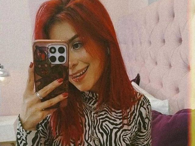 LolyYunno mit roten Haaren, einem zebra-gemusterten Hemd und roten Nägeln, lächelt in die Kamera und hält ein Handy mit einer rosa Hülle, wobei ihre Zähne zu sehen sind. Die Brüste sind nicht sichtbar..