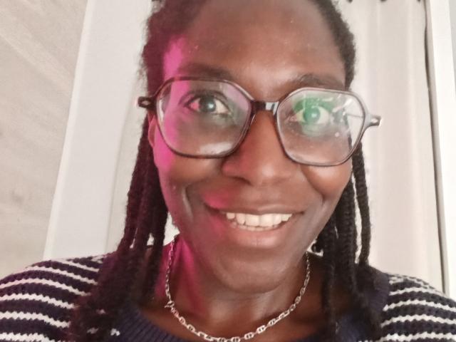 NerySkiraa has dark skin, dreadlocks, and wears glasses with a reflection of green eyes. She's smiling with white teeth, wearing a striped shirt and a silver chain.