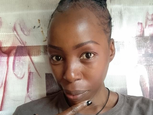 A BeutyLicious tem uma pele marrom brilhante, olhos escuros e um sorriso discreto. Ela está tocando o queixo com unhas pintadas de preto, vestindo uma camisa cinza e tem o cabelo trançado..