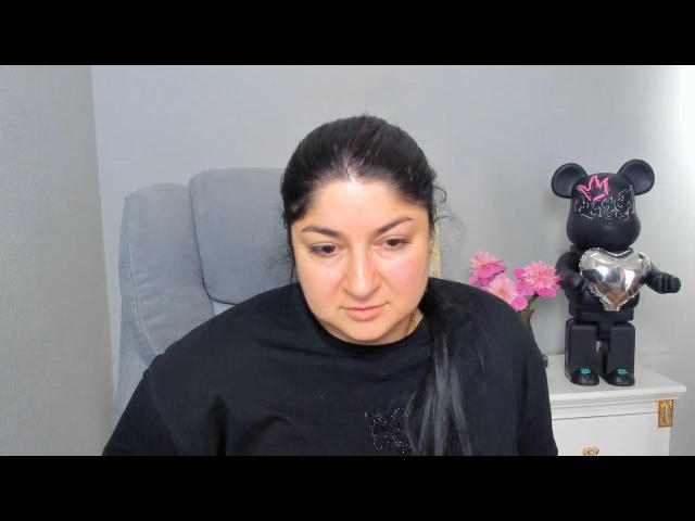 Candenlin has a chubby face, dark hair in a ponytail, wearing a black shirt, sitting in a gray chair, with a black bear statue and pink flowers in the background.