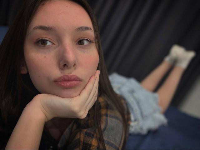 AnthonyCellio liegt auf einem Bett, das Gesicht auf der Hand, braunes Haar, rosafarbene Haut, kariertes Hemd, Jeansshorts, weiße Socken, leicht geschwollene Lippen, braune Augen, unscharfer Hintergrund..