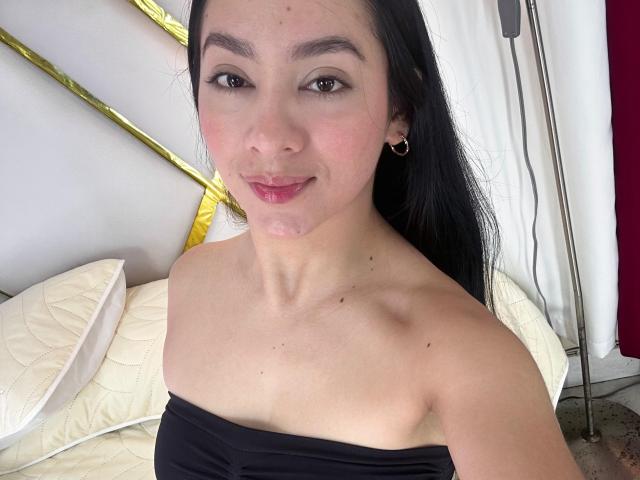 LilaPink mit schwarzen Haaren, kleinen Brüsten, freiliegenden Schultern, einem schwarzen Top mit Trägern, einem leichten Lächeln, rosafarbenen Lippen, heller Haut, kleinen Ohrringen, sitzend auf einem Bett mit weißen Kissen..