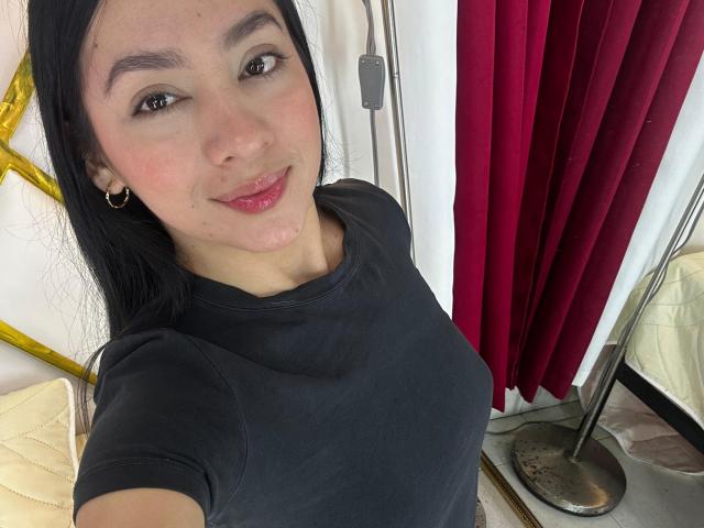 LilaPink con una camisa negra, pechos pequeños, ojos marrones, labios rosados, cabello negro, aretes dorados, sonriendo, una cortina roja, una pared blanca y un espejo con marco dorado en el fondo..