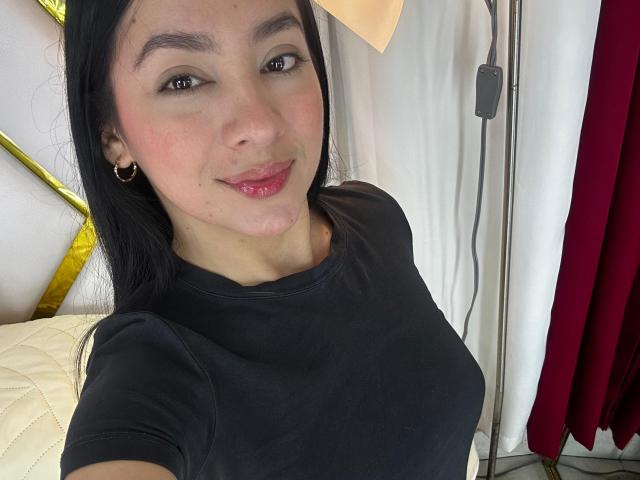 LilaPink con capelli neri, occhi marroni, rossetto rosa, piccoli orecchini, camicia nera, sorridente, guancia leggermente gonfia e rossa, letto con cuscini bianchi e gialli, tenda rossa, lampada..