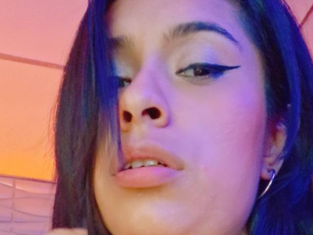 RoxanneHan with dark hair, bold black eyeliner, slightly parted lips, and a small pimple on her right cheek. Blue and pink lighting highlights her face.