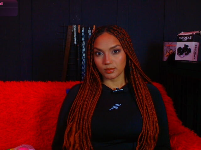 BrianaPratt with long red braids, wearing a black shirt, sitting on a red fluffy couch, has a nose ring, and a sexy, confident expression.
