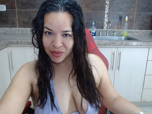 LizethBells, com cabelo preto molhado, vestindo uma blusa roxa com decote baixo, mostrando seus seios grandes, sentada em uma cadeira vermelha em uma cozinha, sorrindo levemente, com batom vermelho..
