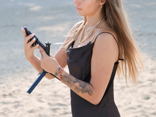 AmmaLoyXX with long blonde hair, black tank top, tattooed arm, holding blue flashlight, wearing gold necklace, standing on sandy beach, focused on flashlight.