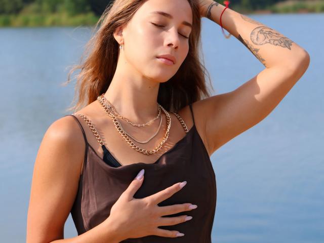 AmmaLoyXX with tan skin, brown hair, and tattoos, wearing a brown top with gold chains, eyes closed, hand on chest, other hand in hair, by a blue lake.