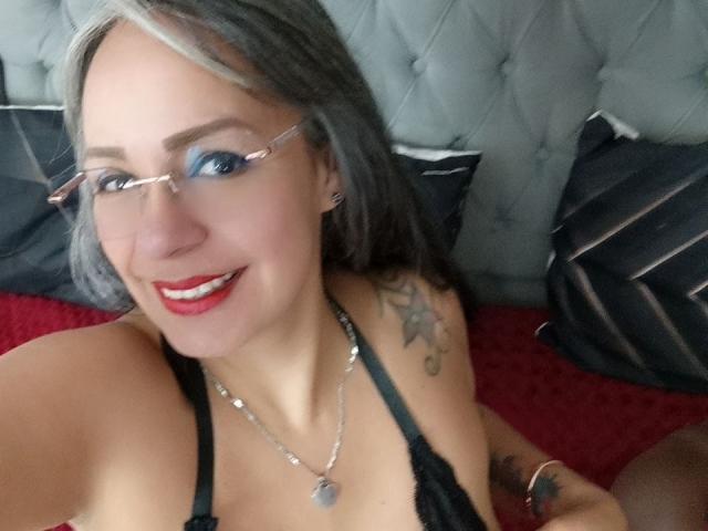 Laila Castillo mit Brille, rotem Lippenstift, schwarzem Spitzenoberteil, grauen Haaren, Tattoos an der Schulter, einer Halskette, lächelnd, rohes Bett, graue Kopfteil. Brüste sichtbar, Nippel durch das Spitzenmaterial hindurch..