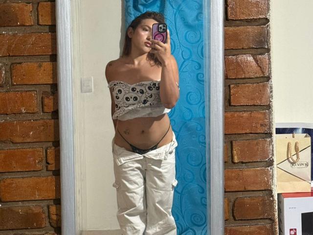 TommyTS, con cabello castaño, pechos pequeños, vistiendo un top de encaje blanco y pantalones blancos bajados para revelar ropa interior negra, tomando una selfie frente al espejo con un teléfono morado..
