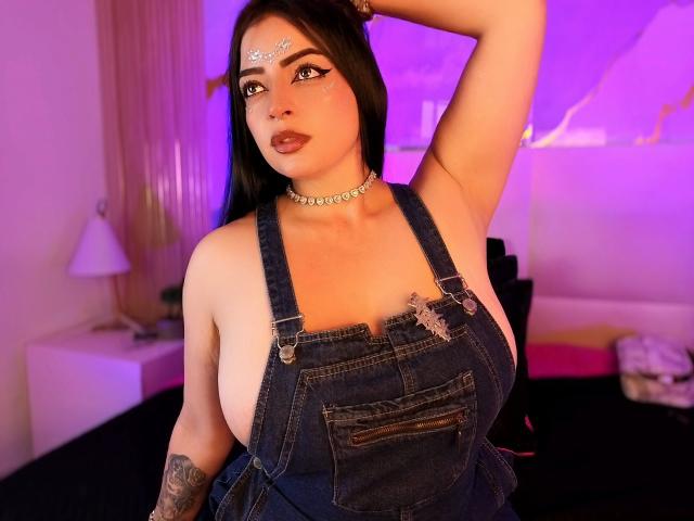 LilithAmoth with huge tits, wearing blue denim overalls, choker, and forehead gem, arm raised, tattoo on left arm, black hair, purple and pink lighting, bedroom setting.
