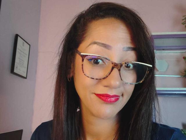 SamanthaCorine ha i capelli castani, occhiali con montatura tartarugata e rossetto rosso. Sorride leggermente, con la testa leggermente inclinata, e indossa una camicia scura..