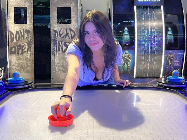 MaraCortes, avec de grandes seins, portant une chemise bleue, penchée sur une machine de flipper, atteignant une bille rouge, souriante, cheveux bruns, dans une salle d'arcade faiblement éclairée..