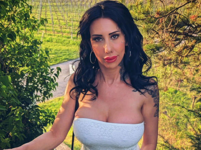 CandySuck, une femme séduisante avec de grandes seins, portant une robe blanche sans bretelles, du rouge à lèvres rouge, des boucles d'oreilles pendantes et des cheveux noirs ondulés, debout à l'extérieur avec du feuillage et un vignoble en arrière-plan..