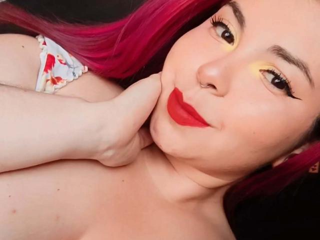 Hada69, avec ses cheveux roses, son eye-liner jaune, son rouge à lèvres rouge, un piercing au nez, et une main sur son menton, exhibe ses seins nus. Sa peau est pâle et son expression est mignonne..