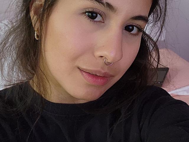Aurora Monroe hat ein süßes Gesicht mit dunkelbraunen Augen, welligem braunem Haar, einem Nasenring und rosafarbenen Lippen. Sie trägt ein schwarzes Hemd und kleine goldene Ohrringe..