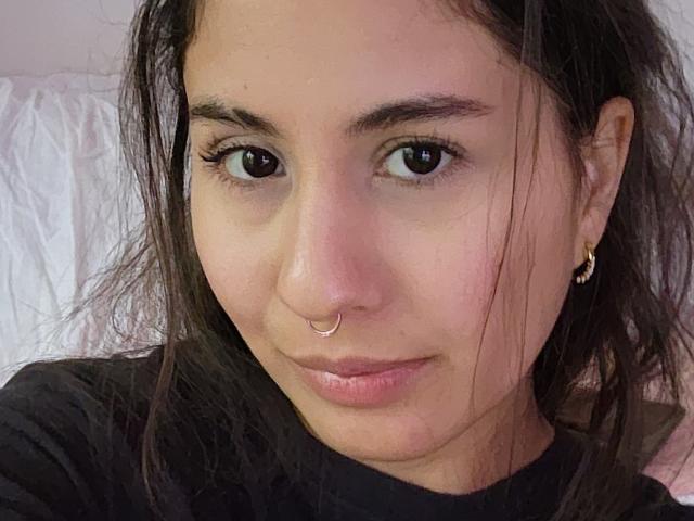 Aurora Monroe, avec un anneau de nez, des sourcils foncés et de longs cheveux noirs, portant une chemise noire, de petites boucles d'oreilles dorées et un sourire discret, montrant son joli visage et ses fesses menues..
