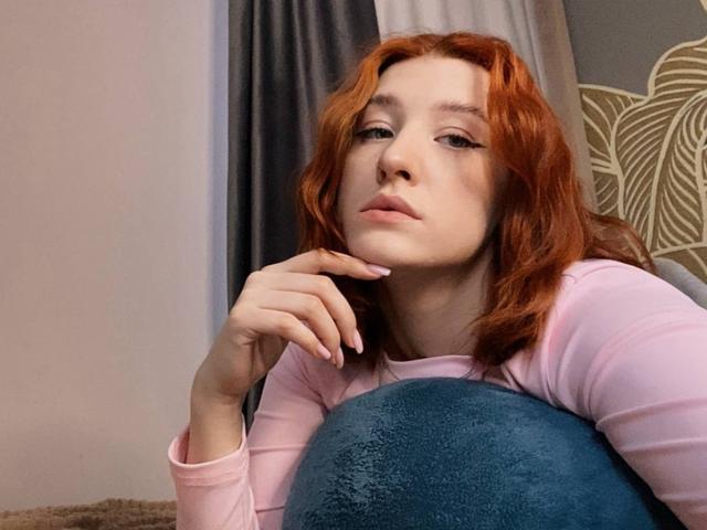 VeolaVanhoecke, con cabello rojo, una blusa rosa y un cojín azul, apoyando el mentón en la mano, con una expresión ligeramente aburrida. Tiene piel clara y delineador de ojos rosa..