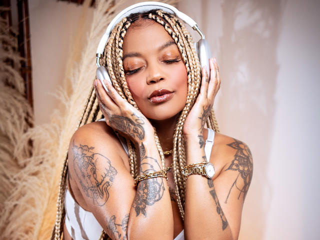 KenyaCarper with braided hair, white headphones, closed eyes, glossy brown lips, tattoos on arms and shoulders, hands on face, gold bracelet, white tank top, soft light.