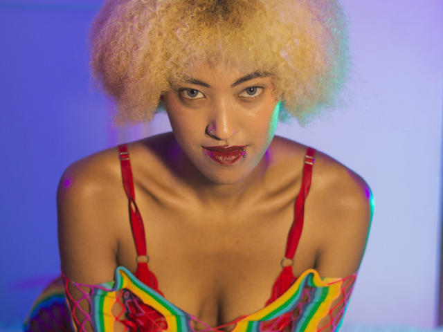 AlishaEbony with curly blonde hair, red bra, and colorful striped top, leaning forward, showing her tits, red lipstick, and nose ring, with blue and purple lighting.