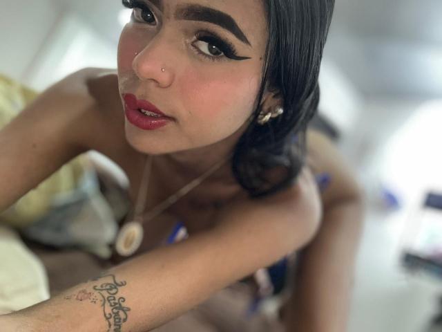 CrystalDavids met donker haar, opvallende eyeliner, rode lipstick en een neusring, onthuld, met een tatoeage op haar arm en een ketting, voorovergebogen met een verleidelijke blik..
