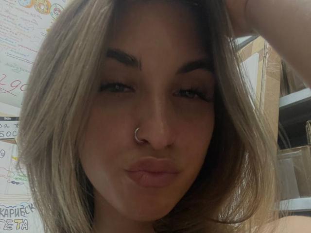 Roxana Krausse, com os lábios curvados, um piercing no nariz e cabelo loiro, olhando para a câmera com os olhos semicerrados, com a mão no cabelo. As nádegas dela não estão visíveis..