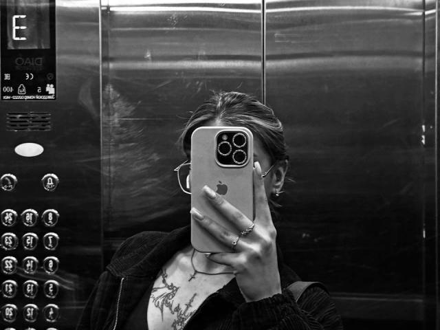 Roxana Krausse tirando uma selfie em um elevador, com o celular cobrindo o rosto, vestindo um casaco, brincos de argola, um colar e um anel, com uma tatuagem visível no peito. Em preto e branco..