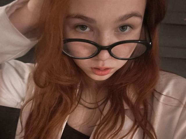 Kati Rokisky mit roten Haaren, schwarzen Brillen und einem weißen Hemd, die mit grünen Augen nach oben blickt, mit leicht geöffneten, rosafarbenen Lippen und einer Hand im Haar. Brüste sind teilweise sichtbar..