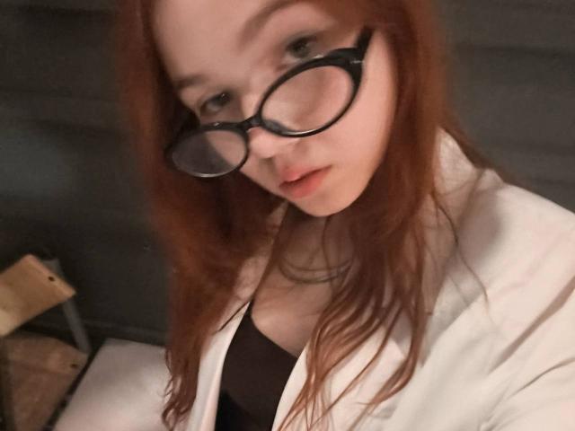 KatiRokisky, avec ses cheveux roux, portant des lunettes noires et un sarrau blanc, lèvres légèrement entrouvertes, montrant ses petites seins, dans un autoportrait flou..