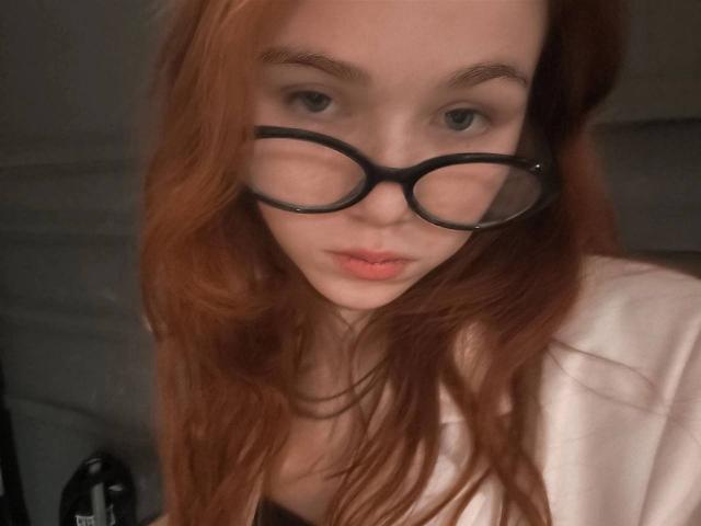 Kati Rokisky mit langen, roten Haaren, die eine schwarze Brille und ein weißes Hemd trägt, blickt direkt in die Kamera mit leicht geöffneten, rosafarbenen Lippen. Ihre Brüste sind nicht sichtbar..