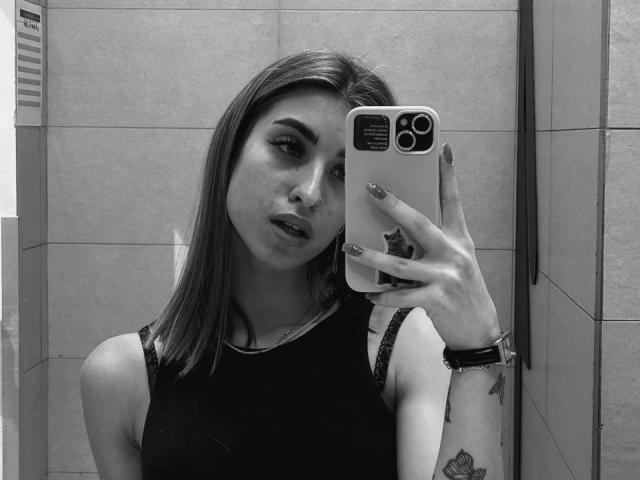 Roxana Krausse che si scatta un selfie allo specchio, mostrando il seno con una canotta nera senza maniche, con lunghi capelli lisci, un tatuaggio sul polso destro, e che tiene in mano un telefono con un pop socket..