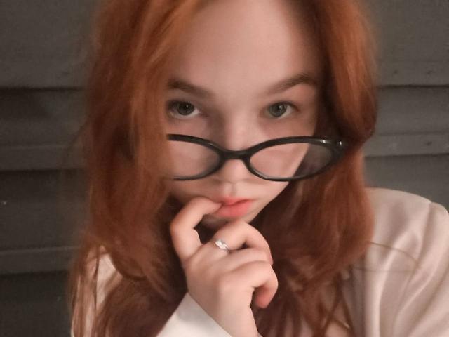 Kati Rokisky, con i capelli rossi, che indossa occhiali neri, che si morde il dito della mano con l'anello, che guarda nella fotocamera, con la pelle pallida e una camicia bianca..