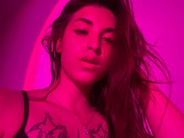 Roxana Krausse, com o cabelo com tons de rosa, com um olhar sedutor, tem tatuagens no peito, veste uma alça preta, seus lábios estão levemente entreabertos e tem uma pequena piercing no nariz..