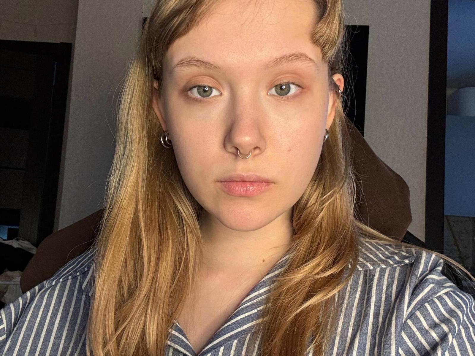 SundayMcgiveron hat hellgrüne Augen, einen Nasenring und einen ernsten Gesichtsausdruck. Sie trägt ein gestreiftes Hemd, und ihr blondes Haar ist offen. Sie schaut direkt in die Kamera..