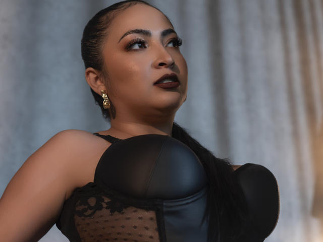 AlanaWest with dark lipstick, wearing black leather bustier, sheer lace, and gold earrings, looking up, braids, large tits, tan skin, sexy, confident, soft lighting.
