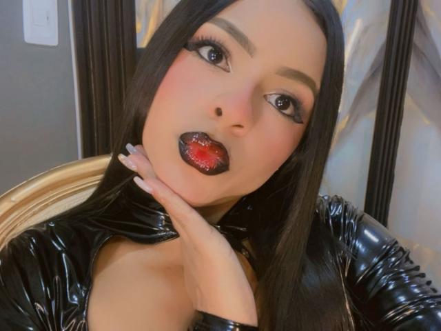 LindaFrancoo vêtue d'une combinaison en latex noir, avec de longs cheveux lisses, du rouge à lèvres foncé, des lèvres rouges, une main sur le cou, de grands yeux, une peau claire, des ongles brillants, regardant la caméra..