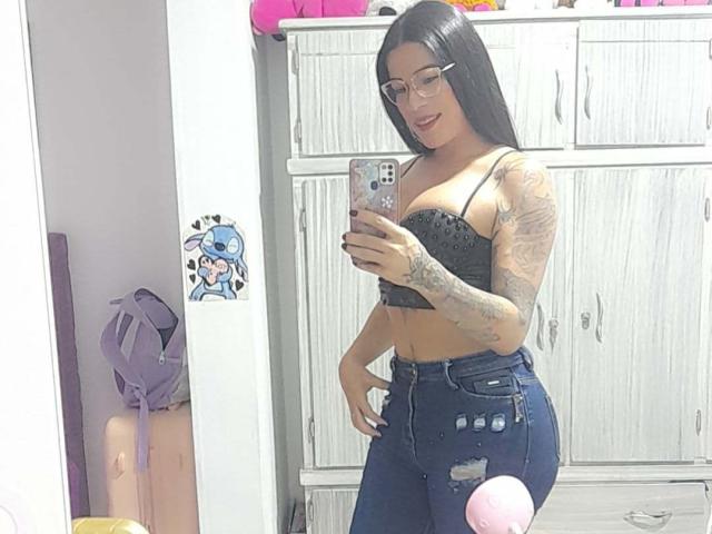 SexyCaandydoll with long black hair, glasses, tattooed arm, black lace bra, and ripped blue jeans, taking a mirror selfie, showing her boobs and cute pink purse.