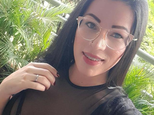 SexyCaandydoll has black hair, glasses, and is smiling with pink lips. She's wearing a sheer black top, showing her tits slightly. Her ringed hand is near her face.