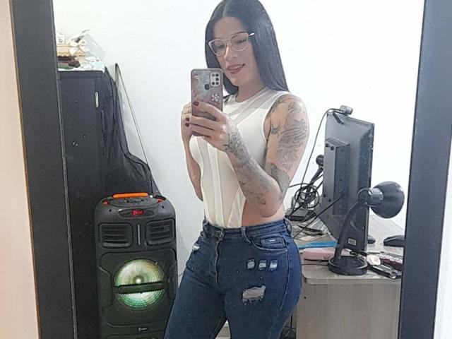 SexyCaandydoll with long black hair, glasses, white sleeveless top, tattoos on arm, wearing tight blue jeans, taking a mirror selfie, standing in front of a computer and speaker.